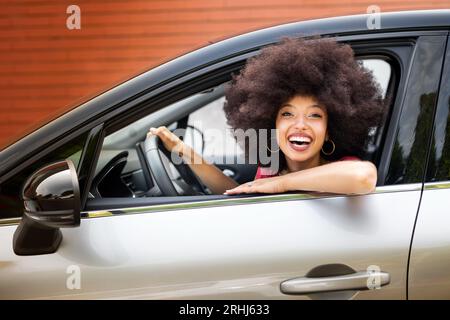 Smiling young African American female with Afro hairstyle looking at camera while sitting in parked car in driver seat with hand on steering wheel nea Stock Photo