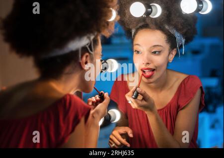 Young Moroccan female with Afro hair headband looking away while standing in front of makeup mirror with illuminated lights and putting red lipstick a Stock Photo