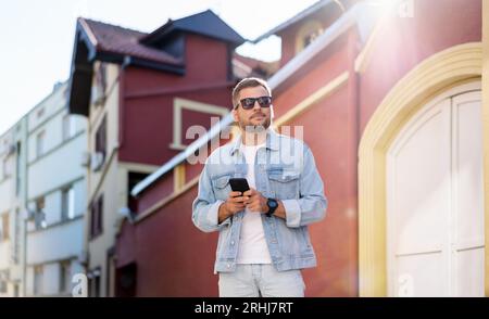 Young adult white caucasian ethnicity man wearing sunglasses and denim jacket standing on city street and using his mobile phone. Stock Photo