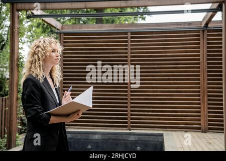 attractive real estate agent holding folder and pen while looking at property on urban street Stock Photo