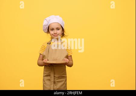 Charming little child, adorable kid girl 6 years old, wearing a chef's hat and beige kitchen apron, smiles cutely and holds out at camera a wooden boa Stock Photo
