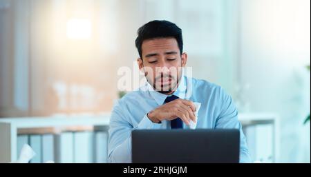 A modern business man feeling sick with covid fatigue while working online at a desk on his laptop. Closeup of an male corporate manager blowing his Stock Photo