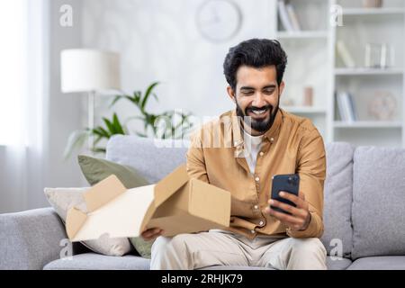 Joyful man at home on the sofa received a package, Latin American is satisfied with the purchase in the online store, writes a positive review using the application on the phone. Stock Photo