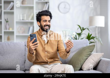 Joyful winner man celebrating victory sitting on sofa at home in living room, hispanic man holding money dollars cash and phone in hands, using smartphone app. Stock Photo