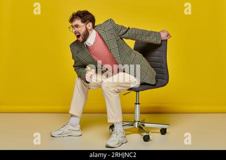 emotional and bearded man screaming and sitting on office chair, yellow backdrop, angry student Stock Photo
