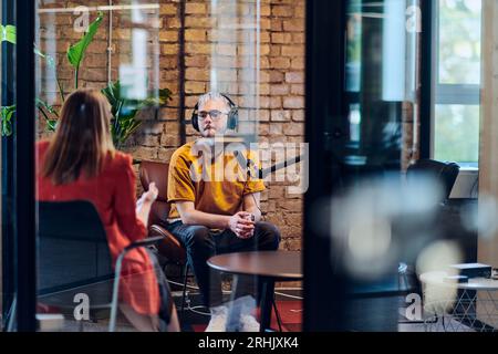 A gathering of young business professionals, some seated in a glass-walled office, engage in a lively conversation and record an online podcast Stock Photo