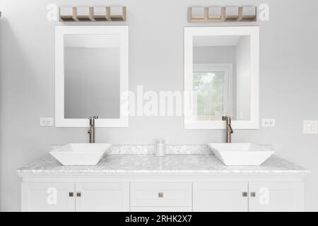 A bathroom detail with a white cabinet, two vessel sinks, marble countertop, and a bronze light fixture mounted above white framed mirrors. Stock Photo