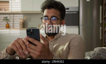 Arabian Indian ethnic man guy use mobile phone on couch browsing mobile internet at home kitchen take break confused read bad news on smartphone male Stock Photo