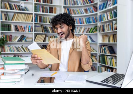 Young student received letter with good exam results and university admission confirmation, hispanic man with curly hair celebrating successful achievement holding hand up close up. Stock Photo