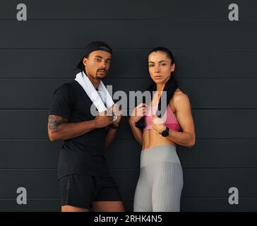 Woman and man in sports clothes at a black wall. Two athletes of different ethnicities holding towels relaxing outdoors. Stock Photo