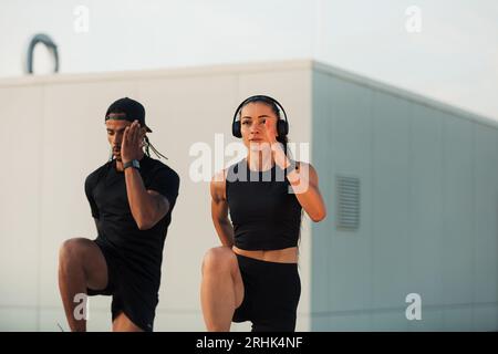 Two athletes doing warming-up exercises on a roof at sunset. Man and woman doing high-intensity exercises together. Stock Photo