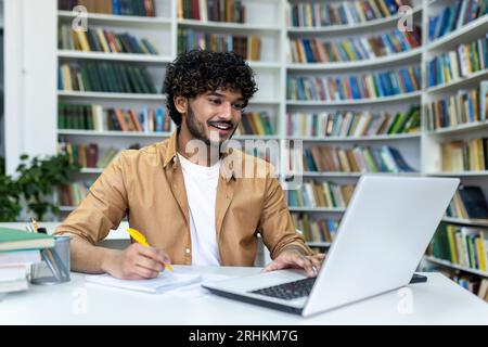 Indian student with curly hair studying sitting among books on shelves, man watching video course writing in notebook smiling contentedly. Stock Photo