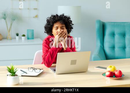 Sleepy african american woman sitting at table with laptop and yawning. Tired overworked girl lazy to work. Student businesswoman relaxing bored in home office after work on laptop computer Stock Photo