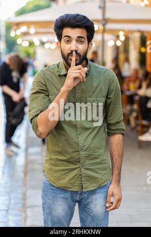 Shh be quiet please. Indian man presses index finger to lips makes silence hush gesture sign do not tells gossip secretoutdoors. Arabian guy walking in urban city street. Town lifestyles. Vertical Stock Photo