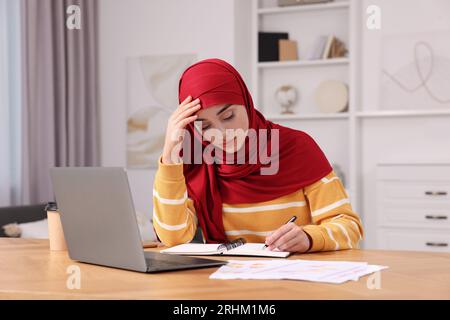 Muslim woman writing notes near laptop at wooden table at home Stock Photo