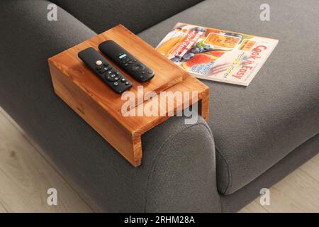Magazine and remote controls on sofa with wooden armrest table in room. Interior element Stock Photo
