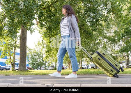 A beautiful woman walks and rolls green suitcases on wheels along the sidewalk. Side view of a happy woman in blue jeans traveling with a large suitca Stock Photo