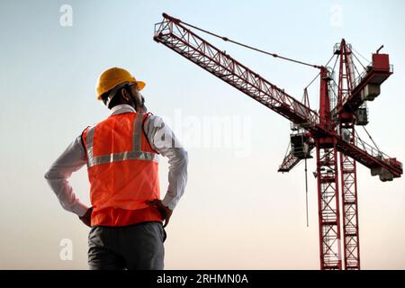Back view of Indian male engineer wearing helmet and vest standing and supervising progress of construction project with crane in the background, Copy Stock Photo