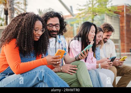 Group of young adult friends using their cellphones and sharing on the social media funny pics outside. Multiracial couple browsing on internet with a phone sitting on a bench with their buddies. High quality photo Stock Photo