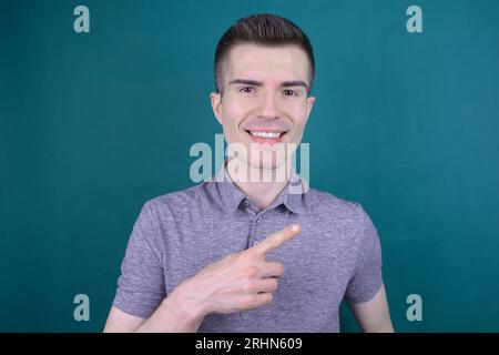 Smiling Young Handsome Caucasian Man in Grey T-Shirt Points His Finger to the Side on Isolated Background Stock Photo