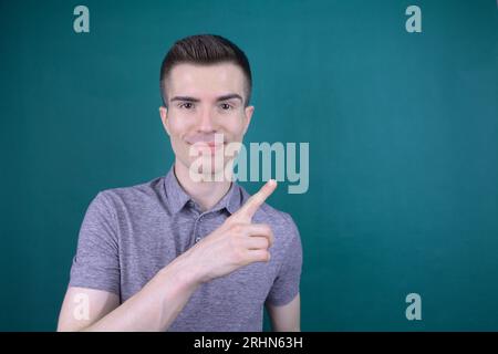 Smiling Young Handsome Caucasian Man in Grey T-Shirt Points His Finger to the Side on Isolated Background Stock Photo