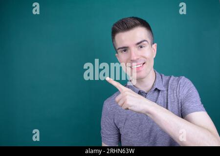 Smiling Young Handsome Caucasian Man in Grey T-Shirt Points His Finger to the Side on Isolated Background Stock Photo