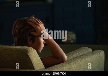 Tired teen boy sitting in green chair with hand on head thinking Stock Photo
