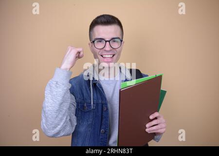 Victorious Handsome Caucasian Student with Backpack, Textbooks, and Books Raises Fist in Triumph on Isolated Backgroud Stock Photo