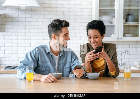 Happy multiracial couple in love, african american woman and caucasian man, using their mobile phones during morning breakfast together at home, watching online videos or news, smile. Using gadgets Stock Photo