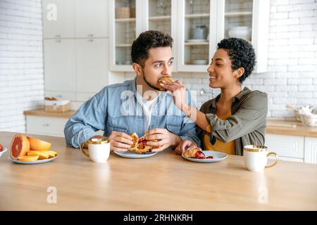 Positive couple enjoying time together. Happy spouses, sit at home in the kitchen, drinking morning coffee with croissants, woman feeding a man a croissant, look at each other, smile Stock Photo