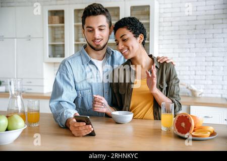 Online video conference. Positive loving couple of different nationalities, talking via video call using smart phone, during a morning breakfast, looking into the camera of the mobile phone, smiling Stock Photo