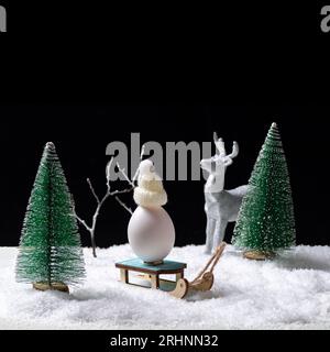 An egg in a white cap sitting on a sled and a toy shining silver deer in a snowy forest among Christmas trees at night. Winter sports. Creative winter Stock Photo