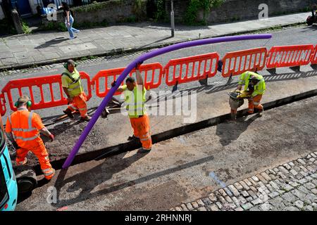 Workmen installing ducting for fibre optic cable in trench cut in road with vibrating whacker compacting bottom of trench before duct is in place, UK Stock Photo