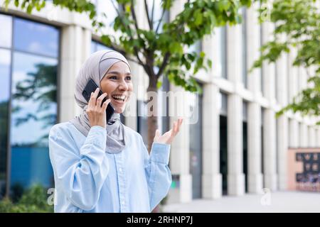 Young joyful businesswoman in hijab talking on the phone outside an office building. Stock Photo