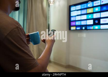 A young guy using smart remote to stream apps on smart TV in house while holding a coffee mug in hand Stock Photo