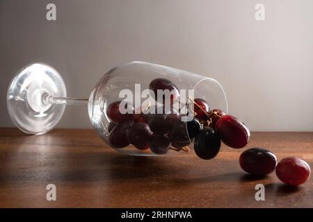 A wine glass falling on wooden table and dark red grapes flowing out against white background Stock Photo