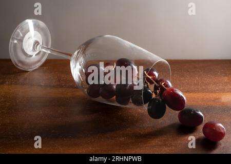 A wine glass falling on wooden table and dark red grapes flowing out against white background Stock Photo