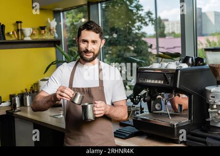 cheerful bearded barista in apron holding pitchers near coffee machine in coffee shop Stock Photo