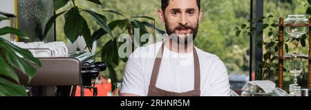 cheerful bearded barista in apron looking at camera, standing and working in coffee shop, banner Stock Photo