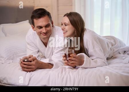 Smiling couple wearing bathrobes and laying in bed with teacups Stock Photo