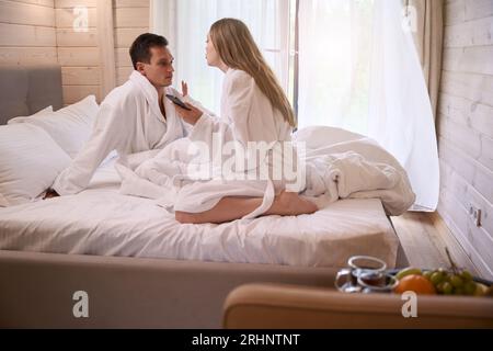 Couple wearing bathrobes and sitting on white sheets in bed Stock Photo