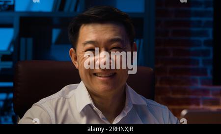 Happy Asian Korean businessman typing laptop working in Internet in office at night evening adult carefree Chinese man leader Japanese male manager Stock Photo