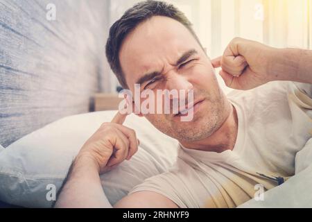 A man at home tries to fall asleep, noisy neighbors interfere with sleep, an guy closes his ears with hands tired after work Stock Photo