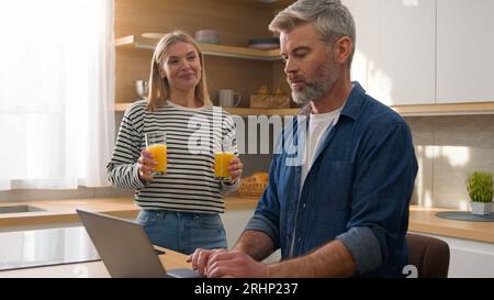 Caucasian adult woman pour orange juice give glass husband man working online with laptop in home kitchen clinking glasses of fresh talking drinking Stock Photo