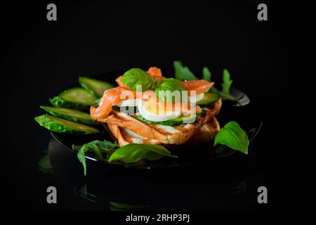 Cooked sandwich with lightly salted red fish, cucumbers and boiled eggs with herbs, isolated on a black background. Stock Photo