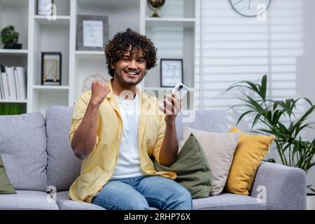 Portrait of young successful man at home on sofa, winner smiling and looking at camera holding phone in hands using game app, holding hand up successful gesture of triumph. Stock Photo