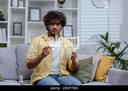Sick man at home with cold and fever, hispanic sitting on sofa in living room holding cup of hot drink with medicine and body thermometer. Stock Photo