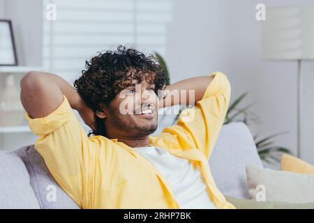 Young man at home on the sofa dreaming smiling and looking out the window, Indian man with his hands behind his head in the living room thinking with a smile. Stock Photo