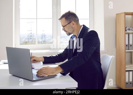 Mature businessman or accountant working in his office, using a laptop, and taking notes Stock Photo