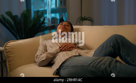 Tired African American woman in evening night living room lying on cozy sofa falling down on comfortable soft couch relaxed tranquil calm girl dream Stock Photo